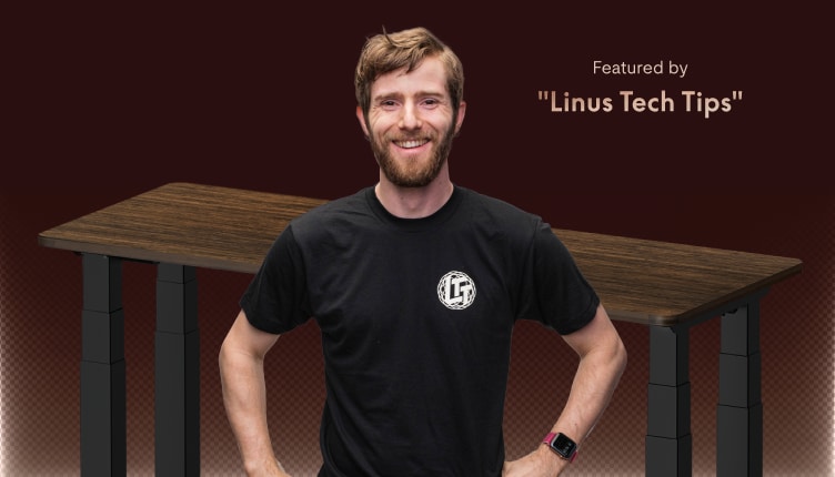 Standing desk with a dark wood finish and adjustable black legs, endorsed by "Linus Tech Tips," featuring a smiling individual in a black T-shirt.